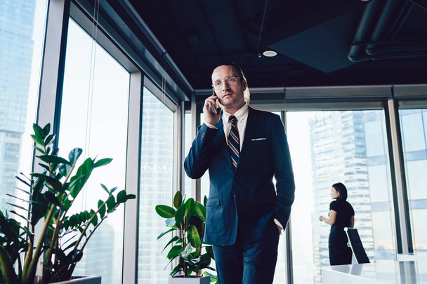 Confident mature man in suit calling via cellular phone while walking in office interior, female colleague standing on background, concept of technology and communication. Male entrepreneur using tech - Photo, image