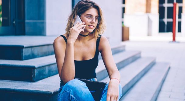 Positive female student communicate with friend after university via smartphone gadget on urban setting, attractive hipster girl calling to friend using mobile application and 4g internet in roaming - Valokuva, kuva