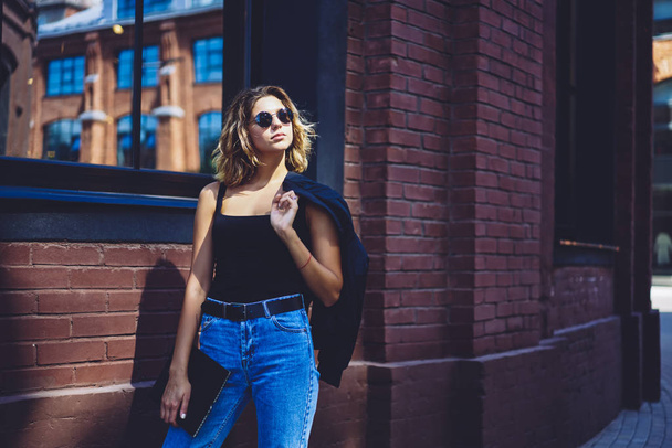 Charming caucasian model in sunglasses looking away and enjoying spring weather while walking on town streets with folder in hands, attractive female student in trendy apparel returning from college - Foto, afbeelding