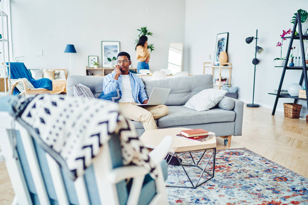 Hombre afroamericano serio freelance hablando por teléfono móvil durante el trabajo a distancia en el interior de la casa moderna, nebuloso tipo hipster desnudo haciendo llamadas mientras se sienta en el sofá en muebles contemporáneos.  - Foto, Imagen