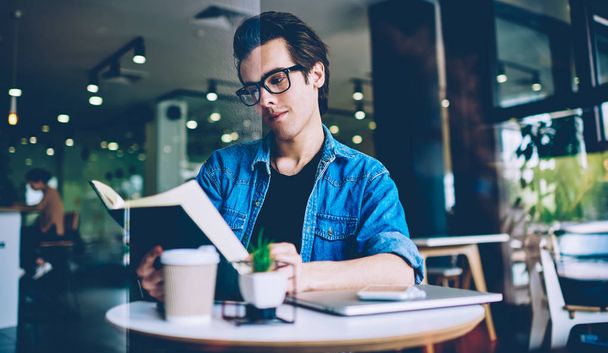 Thoughtful hipster guy in optical spectacles reading knowledge literature in coffee shop for making research of information for course work, pondering man holding textbook for education in hands - Foto, imagen