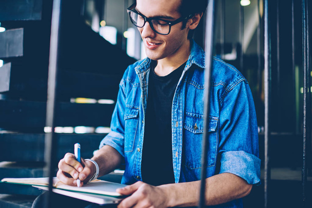 Smiling male student designer drawing sketches in textbook for education during free time on outdoors stairs, positive hipster guy in eyewear writing essay with good idea in notebook on leisure - Φωτογραφία, εικόνα