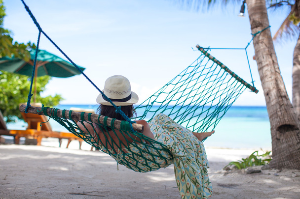 Nainen hatussa makaa riippumatossa puun varjossa rannalla.
 - Valokuva, kuva