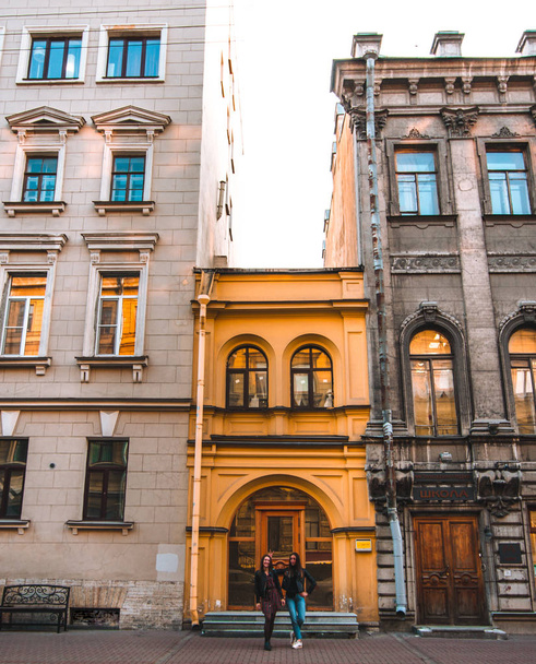 Две привлекательные кавказские девушки стоят рядом с красивыми старыми европейскими зданиями в солнечный день в Санкт-Петербурге. Здание желтого цвета. один друг дает кролику уши над другим
 - Фото, изображение