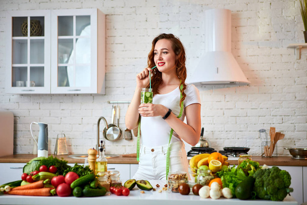 Young woman drinking fresh water with cucumber, lemon and leaves of mint from glass in the kitchen. Healthy Lifestyle and Eating. Health, Beauty, Diet Concept. - Фото, изображение