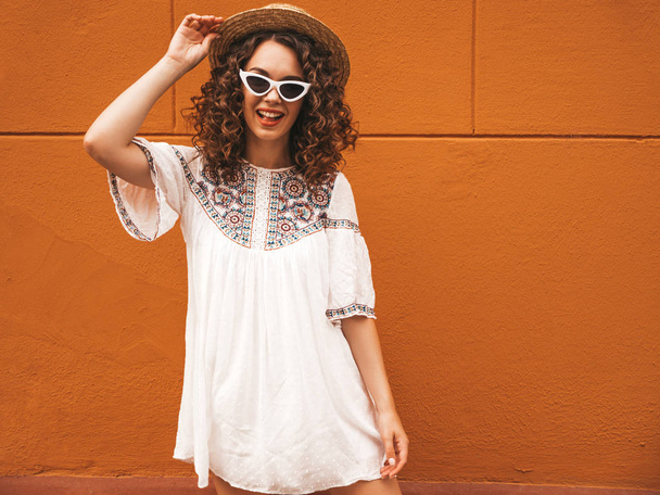 Beautiful smiling model with afro curls hairstyle dressed in summer hipster white dress and sunglasses.Carefree girl posing in the street near yellow wall in hat.Funny and positive woman having fun - Foto, Imagem