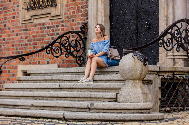 Young woman sitting on a stairs of old house and drinks coffee - Fotoğraf, Görsel