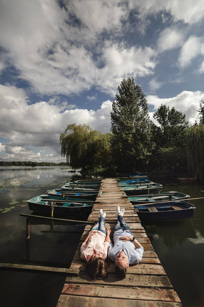 Красивая молодая пара лежит на деревянном мосту на озере, любовно смотрит друг на друга и улыбается. Любовная история. Любящая пара
. - Фото, изображение