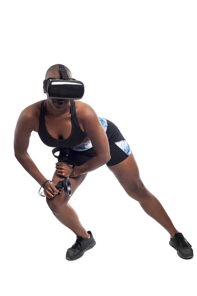 Czarna kobieta nosi zestaw słuchawkowy wirtualnej rzeczywistości i trzyma kontrolery różdżki do ćwiczeń fitness VR. Gracz jest w symulacji sportowej gry wideo dla rozrywki i zdrowej aktywności fizycznej.  - Zdjęcie, obraz
