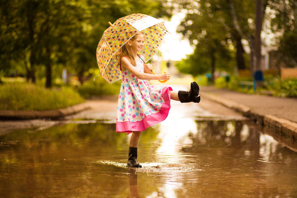Feliz niña divertida con paraguas saltando sobre charcos en botas de goma y en vestido de lunares y riendo
 - Foto, Imagen