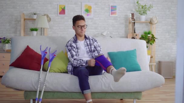 young asian man sitting on the couch puts a tightening elastic bandage on a sick knee - Materiał filmowy, wideo