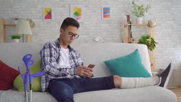 Sad asian young man with a broken leg in bandages, lying on the couch uses smartphone and looking at the camera - Video