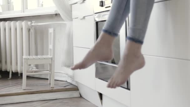 Close up per gambe da donna penzoloni con cassetti da cucina bianchi e forno sullo sfondo. Azione. Donne che indossano jeans seduti a piedi nudi sul bancone della cucina
. - Filmati, video