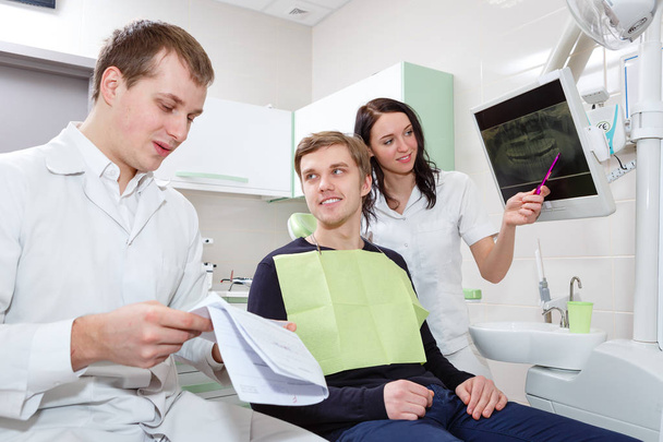 Miespotilas ja hammaslääkäri avustajan kanssa hammashoitolassa tehtävän hammastarkastuksen aikana
 - Valokuva, kuva
