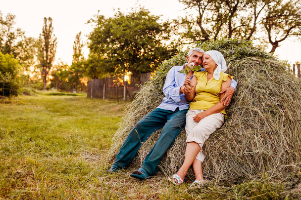 Семейная пара фермеров сидят на стоге сена и отдыхают на закате в сельской местности. Трудолюбивые люди обнимаются
 - Фото, изображение