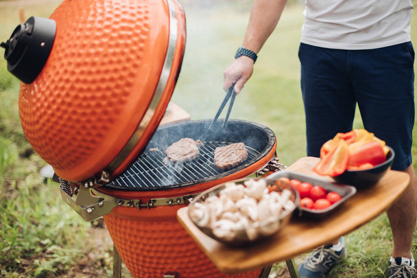 Onherkenbaar man koken vlees op grill. Tafel met gesneden champignons en groenten. Zomer picknick feest. - Foto, afbeelding