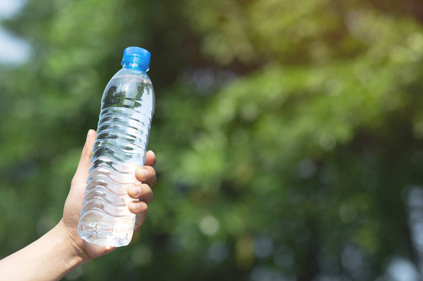 młody człowiek ręka trzyma świeżą butelkę wody pitnej z tworzywa sztucznego w parku. - Zdjęcie, obraz