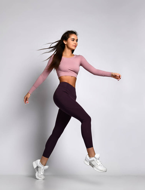 Young athletic woman brunette girl in a good shape with hair in a ponytail in trendy  sportswear gym uniform jumps dances - Foto, imagen