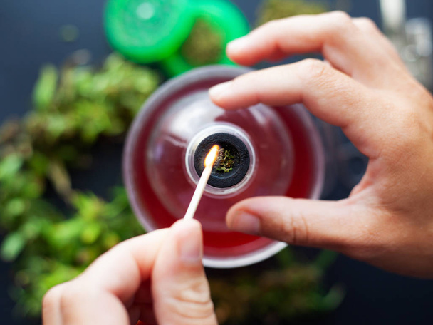 Une main de fille met le feu à un match avec une forte herbe de marijuana. fumer du cannabis dans une bouteille et de l'eau de rose
  - Photo, image