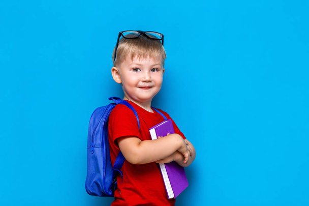 kleiner glücklich lächelnder Junge mit Brille auf dem Kopf, Buch in den Händen, Schultasche auf den Schultern. zurück zur Schule. Schulreif - Foto, Bild
