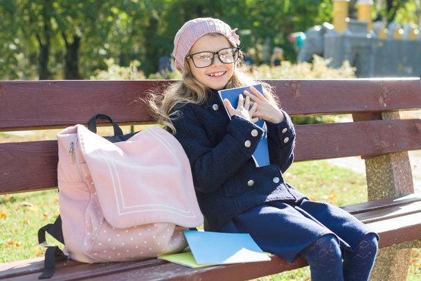 Little girl elementary school student sitting on bench with backpack, looks at school books and notebooks. Background autumn city park - Foto, Imagem