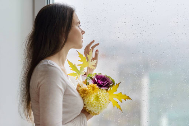 Hello autumn. Young smiling woman with autumn bouquet of yellow and orange flowers, purple ornamental cabbage and maple leaves. Female near the window with raindrops, copy space. - Foto, afbeelding