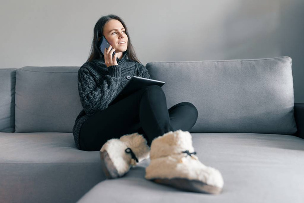 Autumn winter portrait of young girl in warm knitted sweater, at home on the sofa with a digital tablet and phone - Foto, afbeelding