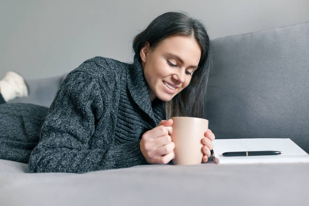 Autumn winter portrait of young girl in warm knitted sweater at home on sofa with notebook and cup of hot drink - Photo, image