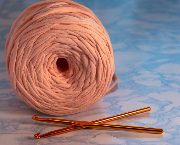 A coil of large knitted yarn of peach color with two orange shiny hooks on the surface of blue marble - Photo, image