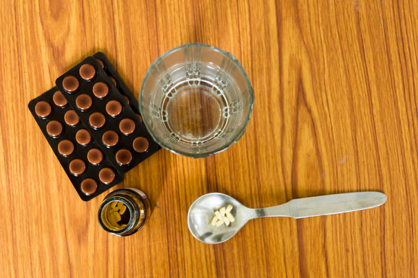 スプーン、ボトル、水のグラスと木製のテーブルの背景にブリスターパックと一緒に薬の錠剤や錠剤のトップビュー。テキスト用コピースペースルームを備えた薬局の治療法とヘルスケア製品. - 写真・画像