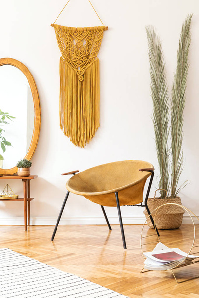 Stylish and minimalistic scandi interior of living room with design gold armchair, lamp, poster frames. dressing table with mirror, plants, palm leaves, yellow macrame and accessories. Cozy home decor - Фото, зображення