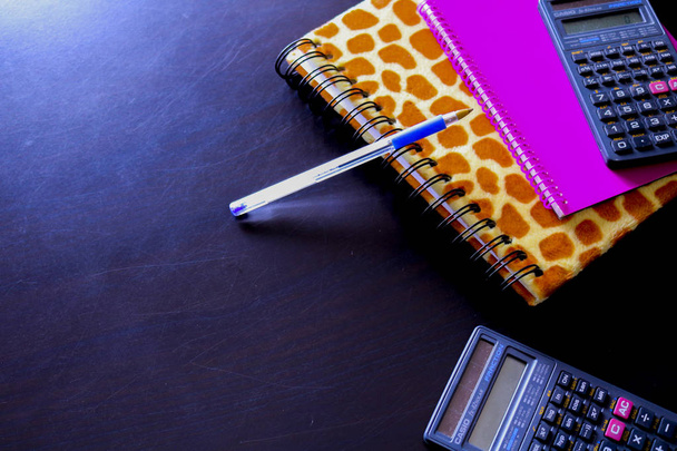 zwei Notizbücher, zwei Taschenrechner und ein blauer Stift mit schwarzem Hintergrund von oben betrachtet, Büroobjekte - Foto, Bild