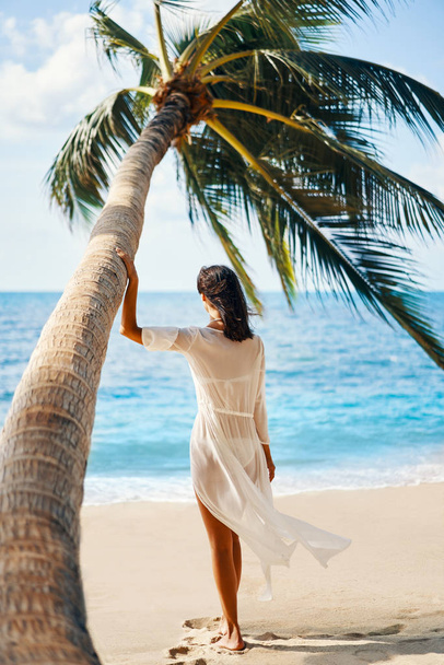 Back view of free young woman enjoying sea standing under palm tree on tropical beach. - Fotoğraf, Görsel