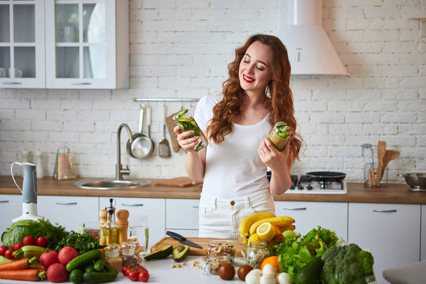 Mujer joven bebiendo batido verde y agua dulce con pepino, limón, hojas de menta en la mesa de la cocina con frutas y verduras. Concepto de alimentación saludable. Menú de comida vegana y desintoxicación
 - Foto, imagen