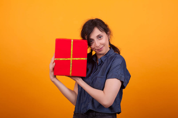Портрет счастливой молодой женщины с красной подарочной коробкой, смотрящей на камеру в студии на жёлтом фоне
 - Фото, изображение