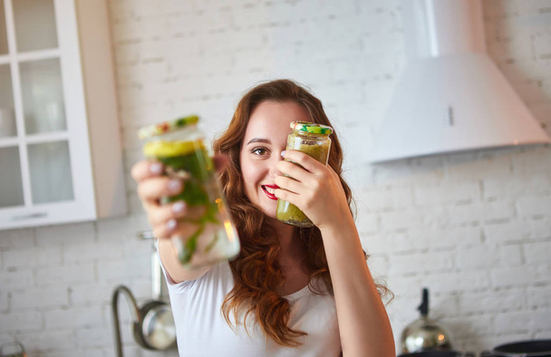緑のスムージーと新鮮な水をキュウリ、レモン、果物や野菜と台所のテーブルの上にミントの葉を飲む若い女性。健康的な食事の概念。ビーガンミールとデトックスメニュー - 写真・画像