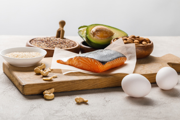 fresh salmon, nuts, eggs and avocado on wooden chopping board near oil isolated on grey, ketogenic diet menu - Photo, Image