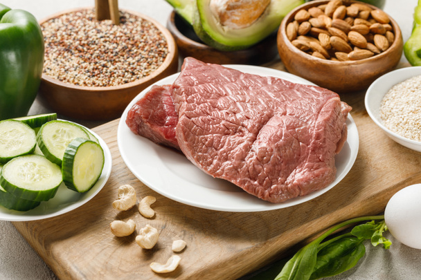 rauw vlees op houten snijplank in de buurt van noten en groene groenten, ketogeen dieetmenu - Foto, afbeelding