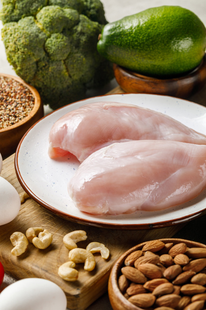 свежие сырые куриные грудки на белой тарелке рядом с орехами, зеленые овощи и яйца, кетогенное меню диеты
 - Фото, изображение