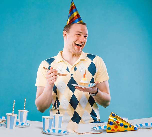 Portrait of man who is celebrating his birthday party, wearing light yellow shirt and party cap on head, making fun, isolated over green background - Photo, Image
