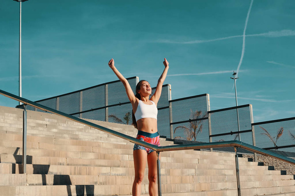 Beautiful young woman in sports clothing keeping arms outstretched while exercising outdoors, standing on stairs  - Photo, image