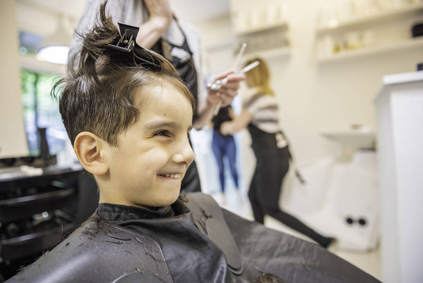 Child little boy in the barber shop hair cut professional smiling portrait looking at mirror - Little boy in the barber shop hair cut professional toddler child getting his first haircut profile - Fotoğraf, Görsel