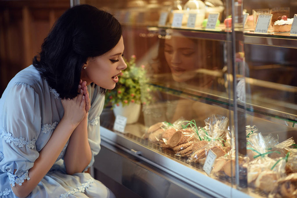 Портрет молодой красивой девушки, смотрящей на витрину с тортами и печеньем с волнением на лице
. - Фото, изображение