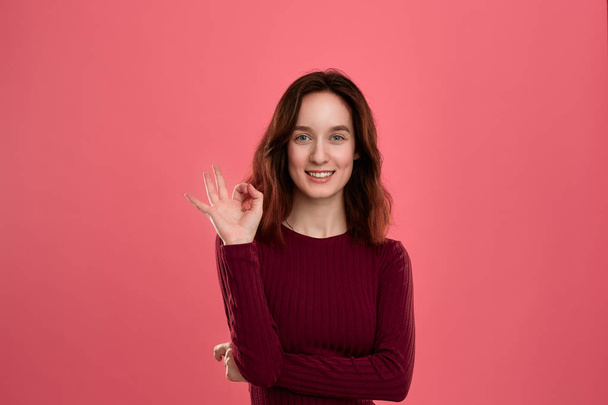 Happy young lady standing isolated over dark pink background looks at the camera while holding palm of a hand near her eye showing gesture of OK-sign. - Fotografie, Obrázek