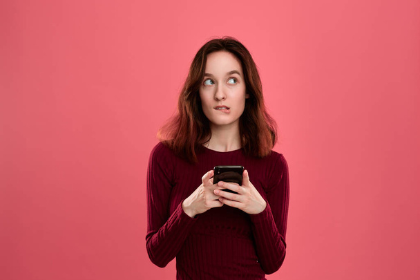 Portrait of a pretty brunette girl standing on a dark pink background and texting on the phone. - Zdjęcie, obraz