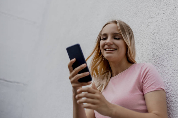 smiling attractive blonde woman using smartphone near grey wall on street - Photo, Image