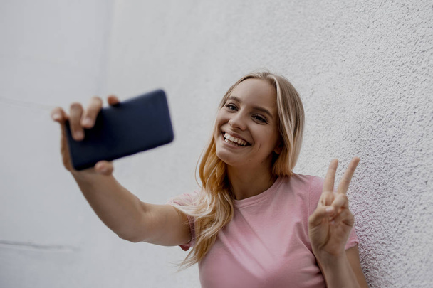 Lächelnde attraktive blonde Frau macht Selfie mit Smartphone in der Nähe einer grauen Mauer auf der Straße - Foto, Bild
