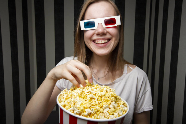 young girl watching a movie and eating popcorn wearing 3d glasses against a striped wall at night, she is emotional and surprised - Foto, Bild