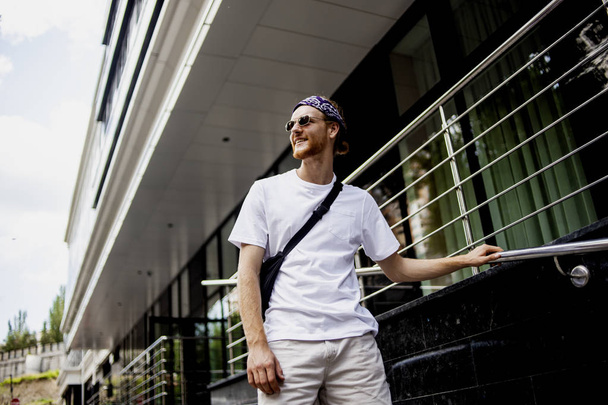 Usměvavý španělský muž v brýlích s zkříženou rukou, oblečený do bílého trička s bílým tričkem a elegantním hipsterem, který prochází městským městem přes černé pozadí - Fotografie, Obrázek