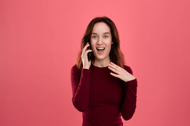 Portrait of a beautiful brunette girl standing on a dark pink background and looking happy while speaking on the phone. - Foto, Imagen